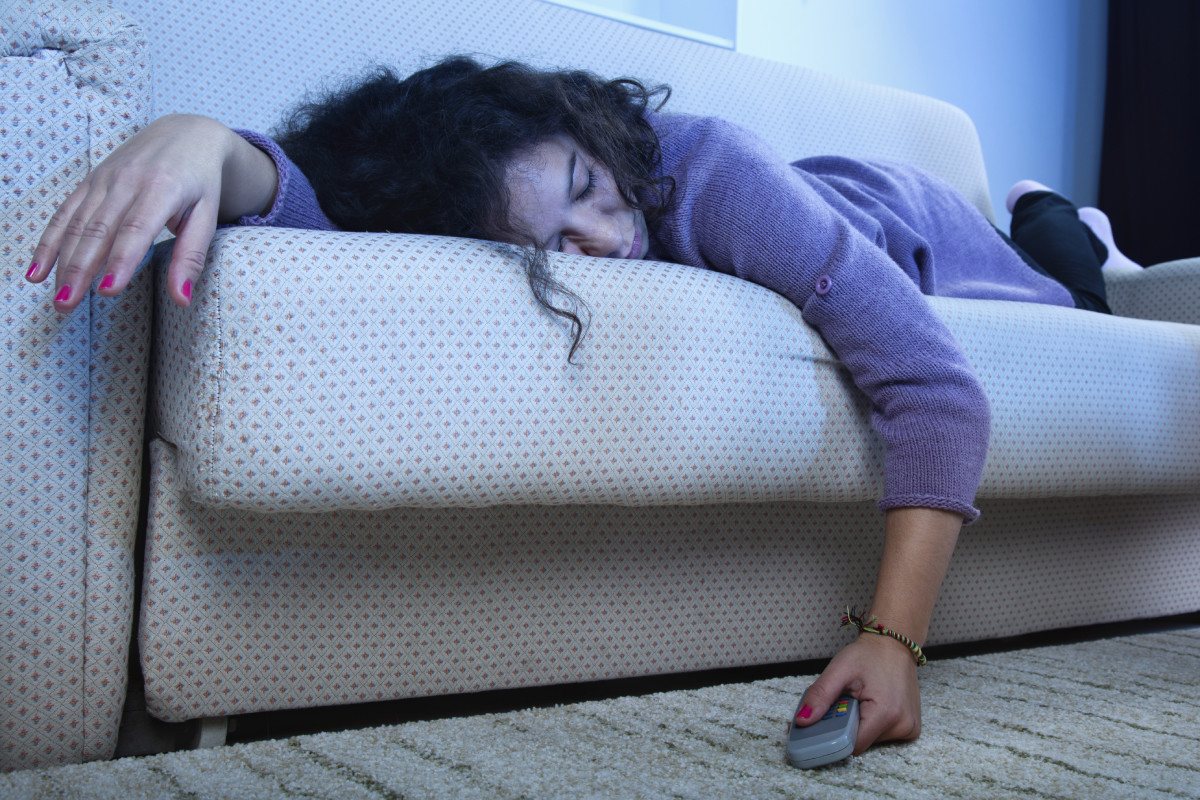 年轻的女人睡在黑暗的客厅里的沙发上，手里拿着电视和遥控器。