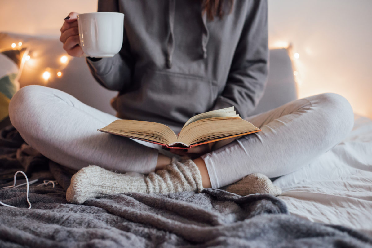 女孩端着一杯热茶，躺在床上看书。她戴着坏耳机，书，智能手机。背景装饰灯。
