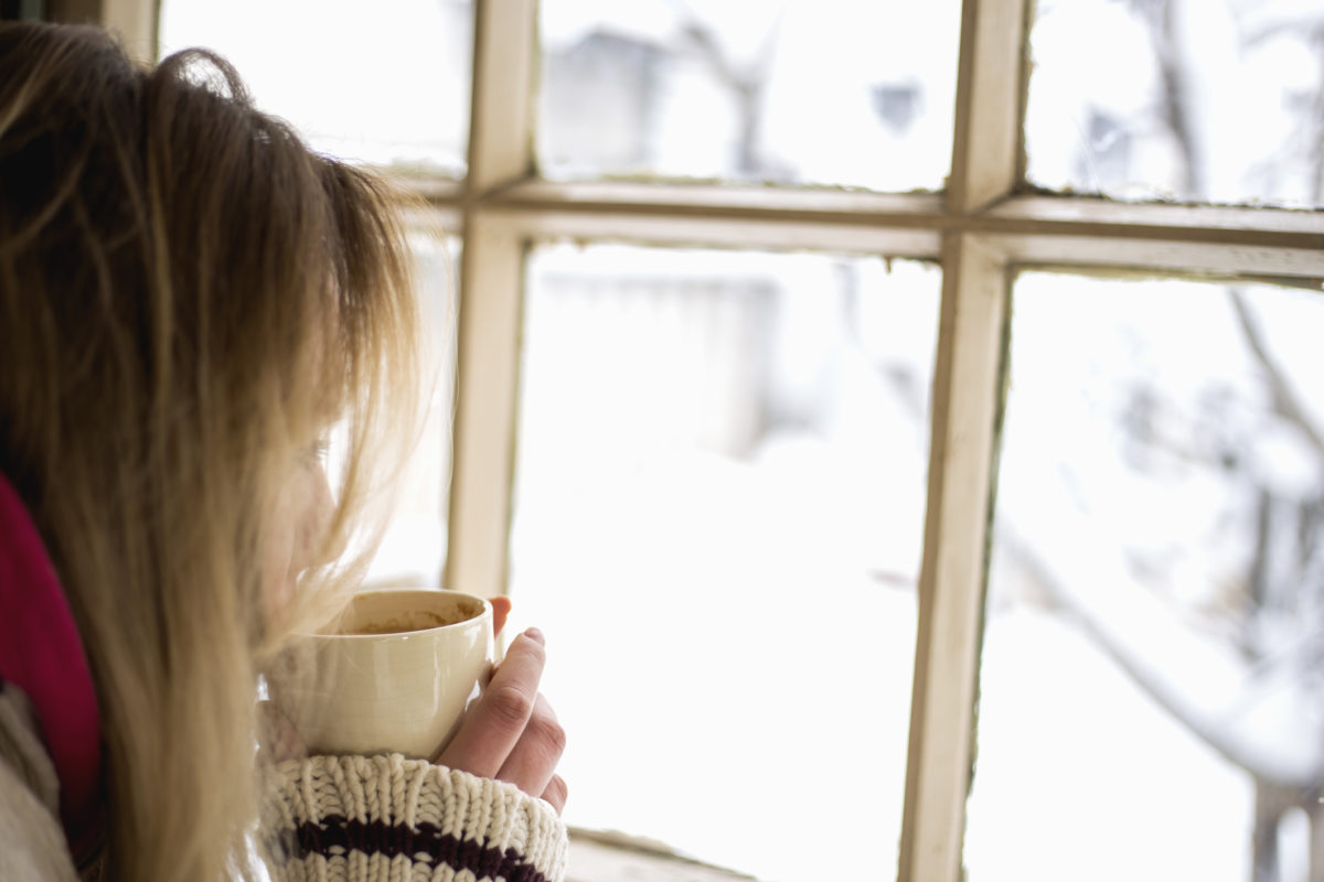 在下雪天喝一杯茶或咖啡的女人的特写镜头。她坐在咖啡馆里透过窗户看。穿着针织开衫和围巾，在温暖舒适的地方过冬。低角度投篮。
