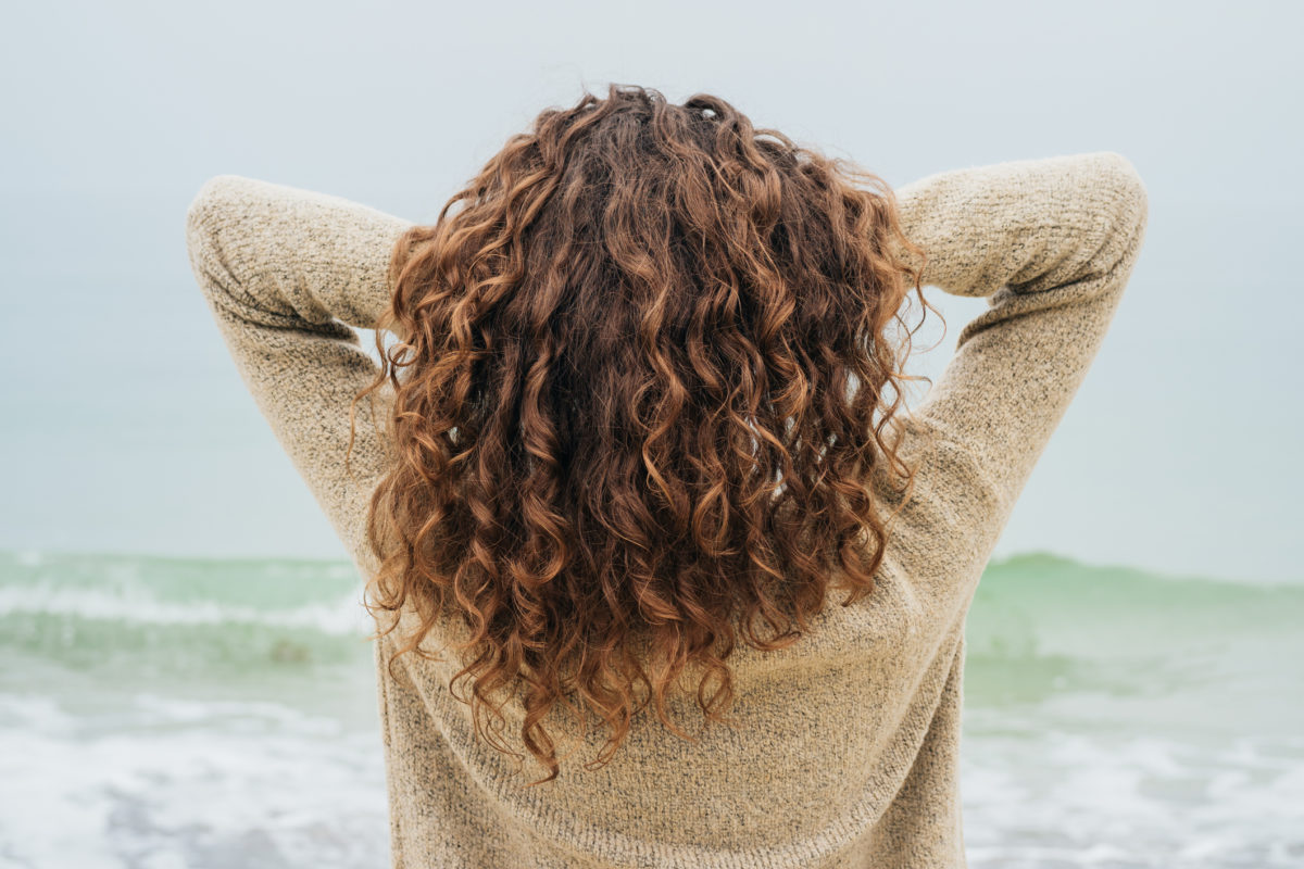 在海边穿着毛衣的卷发黑发女人面向大海，直发，从后面看