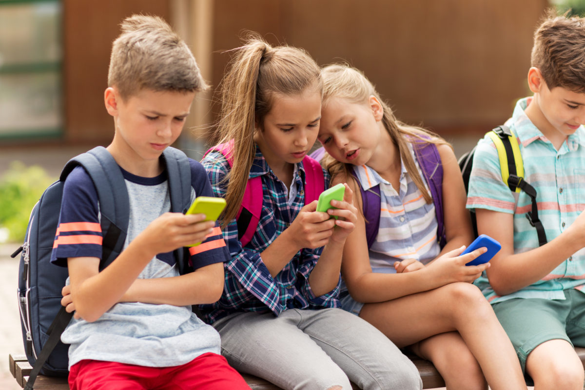 小学教育、友谊、童年、科技和人的概念-一群快乐的小学生，带着智能手机和背包，坐在户外的长凳上