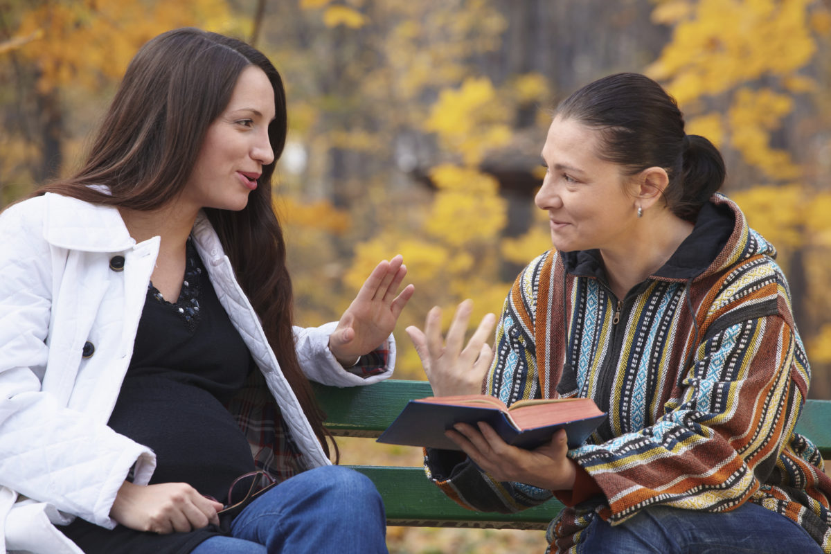 两个女朋友坐在公园里微笑着，一个人正在读圣经中的章节。另一个是晚期妊娠状态。官网下载beplay体育ios版美丽的秋色；真正的人真诚地微笑