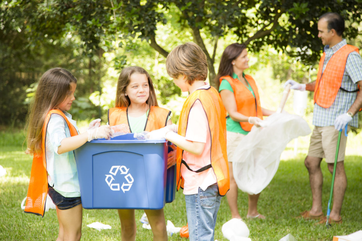 社区志愿者清理他们当地的公园。家庭回收垃圾。孩子们站在前面，手里拿着一个回收箱，爸爸妈妈在后面捡垃圾，手里拿着垃圾袋。的父亲,母亲和三个孩子都穿着安全背心。环境保护。自然。