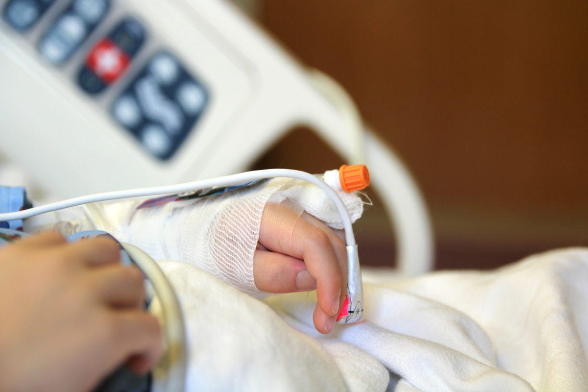 一个孩子躺在医院的床上，手臂和手举过头顶，带着静脉注射。然后用绷带包扎。