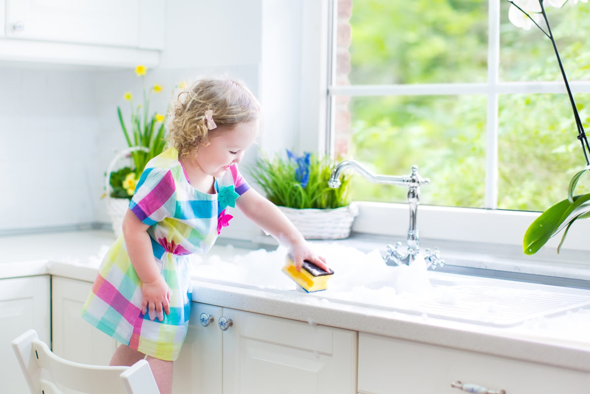 可爱的卷发小女孩，穿着鲜艳的衣服洗盘子，在一个美丽的阳光明媚的白色厨房里用海绵和泡沫在水池里玩，在现代家庭里有一个花园窗