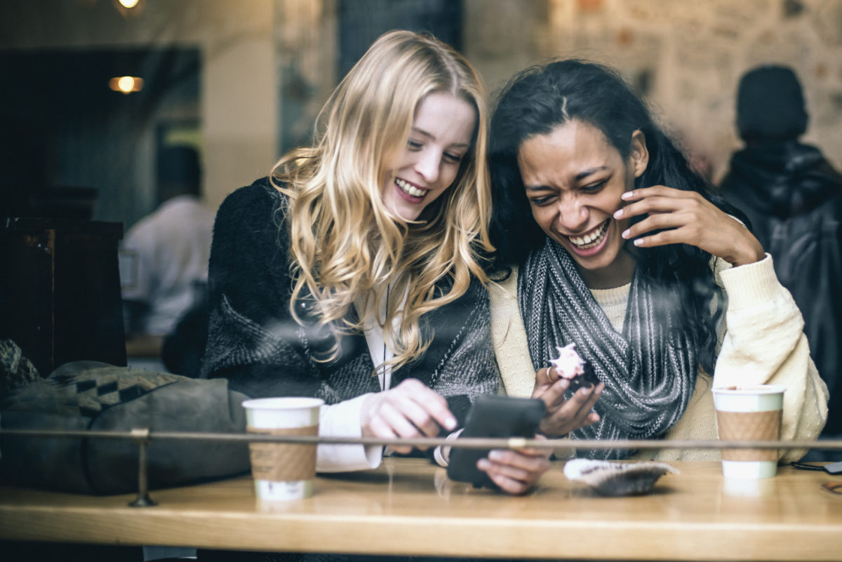 两个多民族的年轻女性朋友玩得很开心，在纽约当地的咖啡店享用咖啡和糕点时，使用智能手机来愉悦回忆，自然光,从街上拿走。