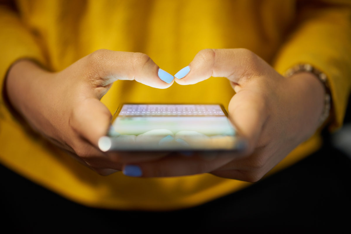 年轻女子晚上用手机在社交网络上发送短信。手的特写与背景中的笔记本电脑
