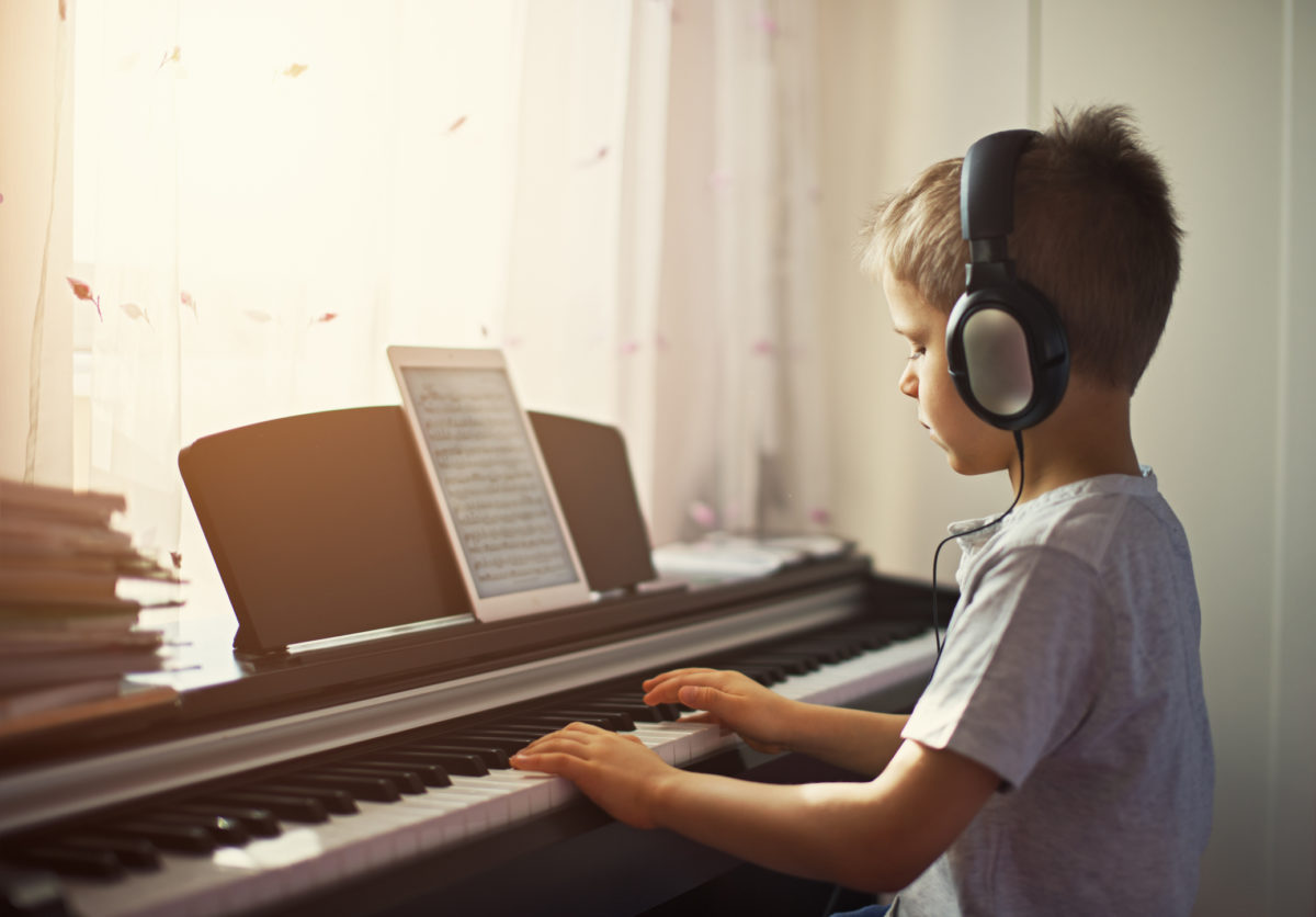 阳光明媚的日子里，小男孩在窗边练习数字钢琴。这个男孩正在使用耳机，平板电脑正在播放音乐。