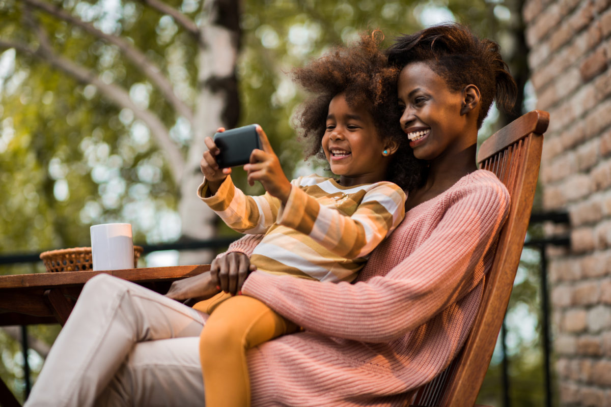 微笑的非裔美国母亲和女儿在阳台上放松，用手机自拍。