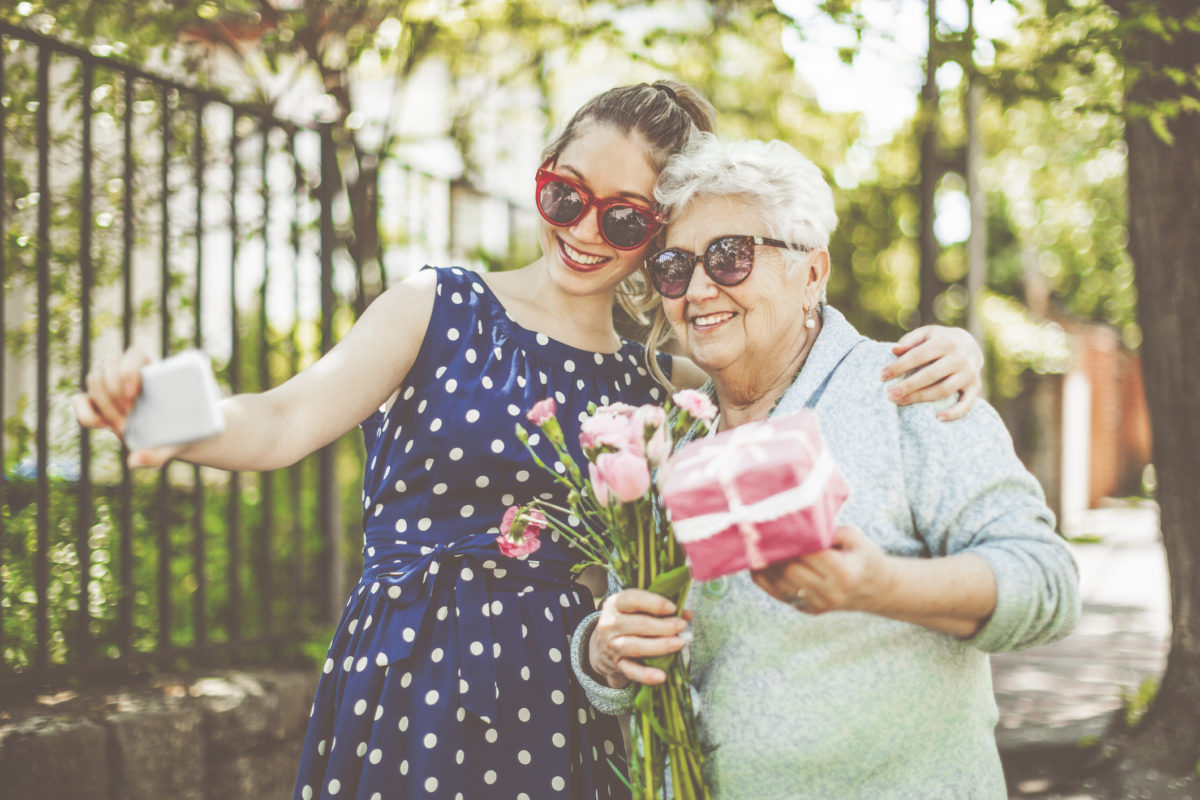 微笑的年轻女孩正在自拍自己和她的祖母，而美丽的老年妇女拿着一份礼物和一束花。