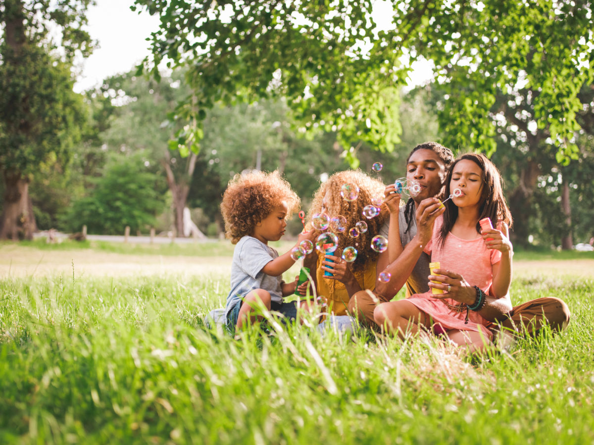 可爱的非裔美国家庭坐在一个美丽的风景如画的公园里，和他们的孩子玩耍，在一个阳光明媚的下午吹泡泡。