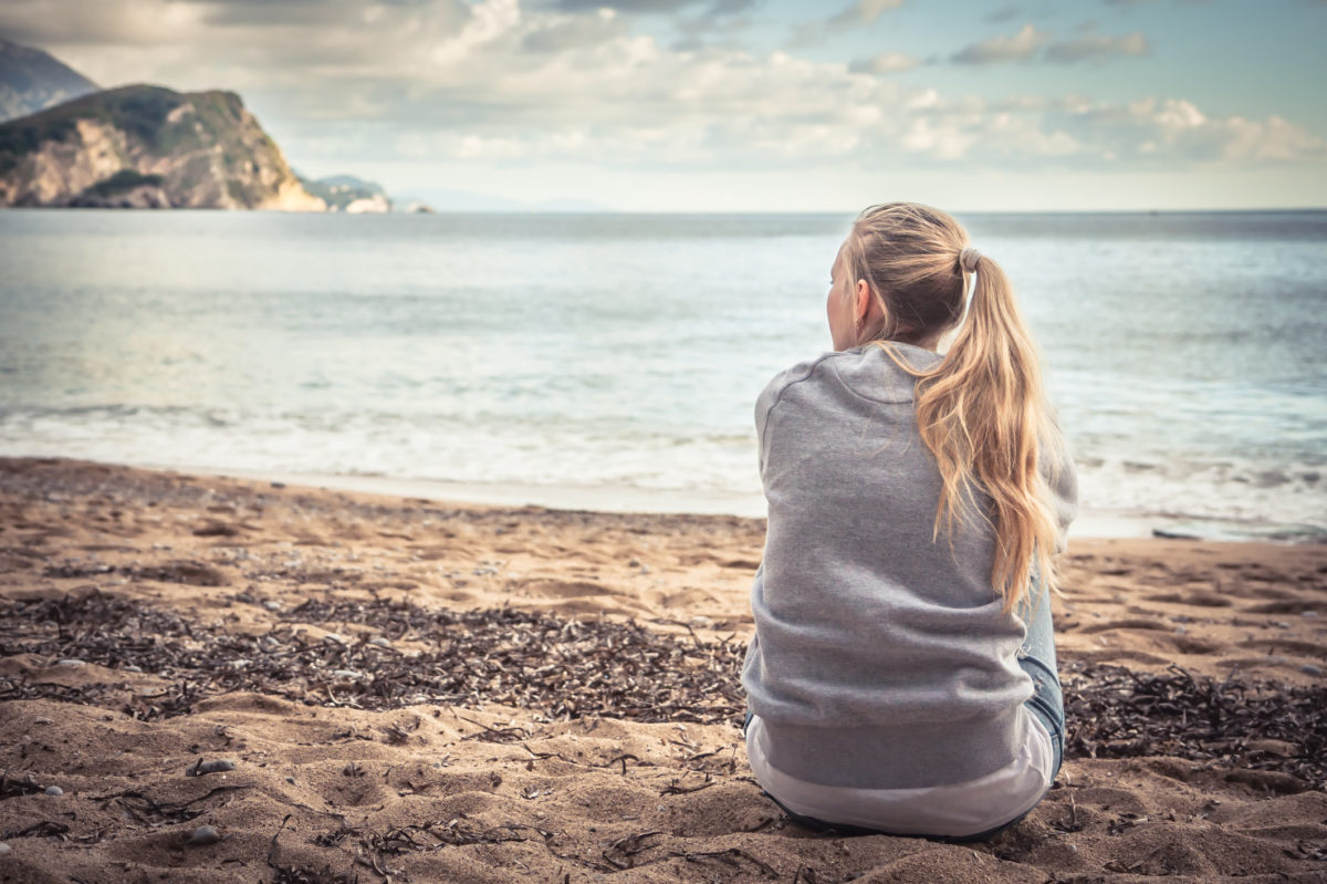 孤单的年轻女游客坐在沙滩上，抱着膝盖，满怀希望地远眺远方。