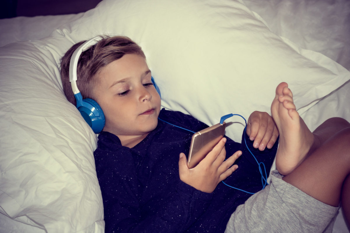 小孩在床上休息，用耳机听音乐。男孩躺在卧室里，用手机看动画片。
